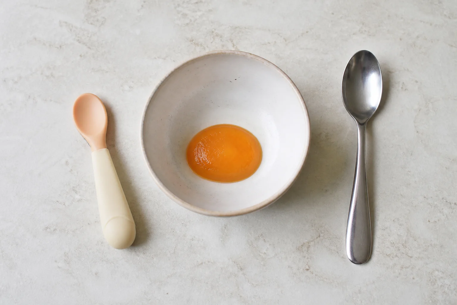 A baby bowl containing a very small two-teaspoon portion of smooth puree beside infant and adult spoons